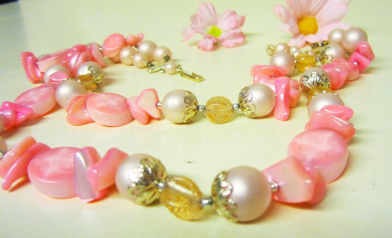 May include: A pink and white beaded necklace with gold accents. The necklace features a variety of pink and white beads, including some with a pearlescent finish. The necklace is strung on a gold chain.