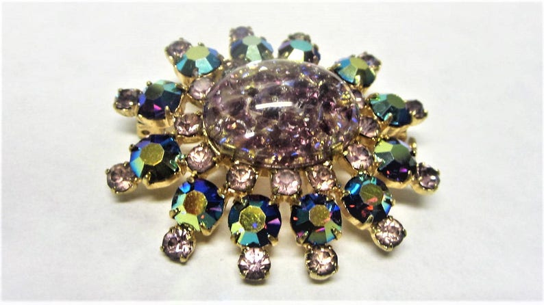 Peut inclure: Une broche dor&eacute;e avec une grande pierre iridescente de couleur violette entour&eacute;e de pierres plus petites, iris&eacute;es, bleues, vertes et violettes.