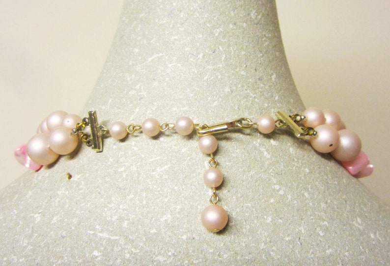 May include: A pink pearl necklace with a gold clasp and a pink flower accent. The necklace is on a white mannequin.