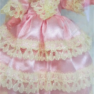 Puede incluir: Un vestido de sat&eacute;n rosa con un ribete de encaje en capas. El vestido tiene un escote y mangas con volantes.