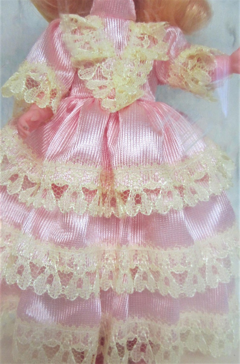 Puede incluir: Un vestido de sat&eacute;n rosa con un ribete de encaje en capas. El vestido tiene un escote y mangas con volantes.
