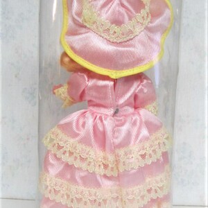 Puede incluir: Una mu&ntilde;eca de porcelana rosa que lleva un vestido rosa con ribetes de encaje y un sombrero rosa con ribetes amarillos. La mu&ntilde;eca est&aacute; en un recipiente de pl&aacute;stico transparente.
