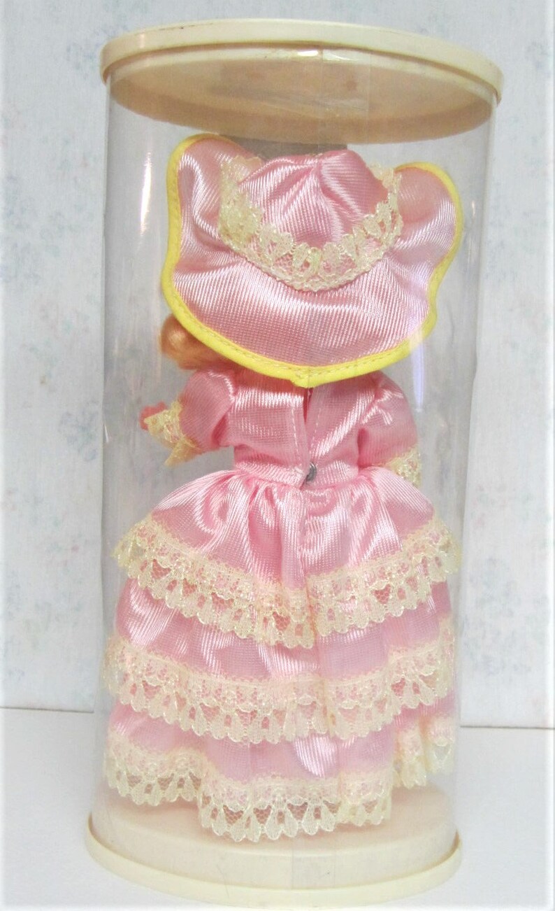 Puede incluir: Una mu&ntilde;eca de porcelana rosa que lleva un vestido rosa con ribetes de encaje y un sombrero rosa con ribetes amarillos. La mu&ntilde;eca est&aacute; en un recipiente de pl&aacute;stico transparente.