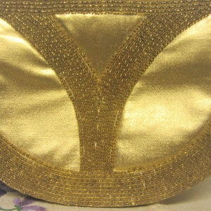 May include: A gold-colored clutch purse with a beaded trim. The purse has a Y-shaped design in the center.