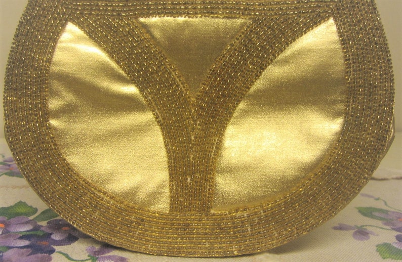 May include: A gold-colored clutch purse with a beaded trim. The purse has a Y-shaped design in the center.