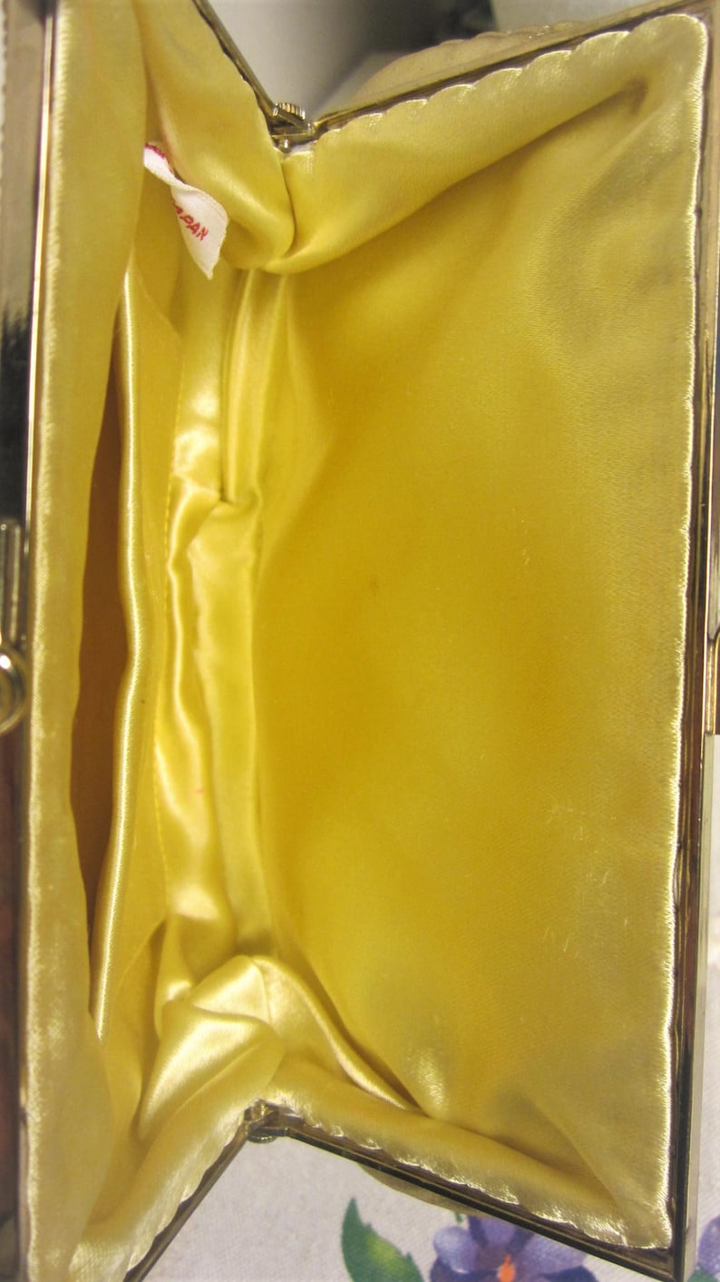 May include: A gold-colored metal purse with a yellow satin lining. The purse is open and the lining is visible. The purse has a small red tag with the word "Japan" printed on it.