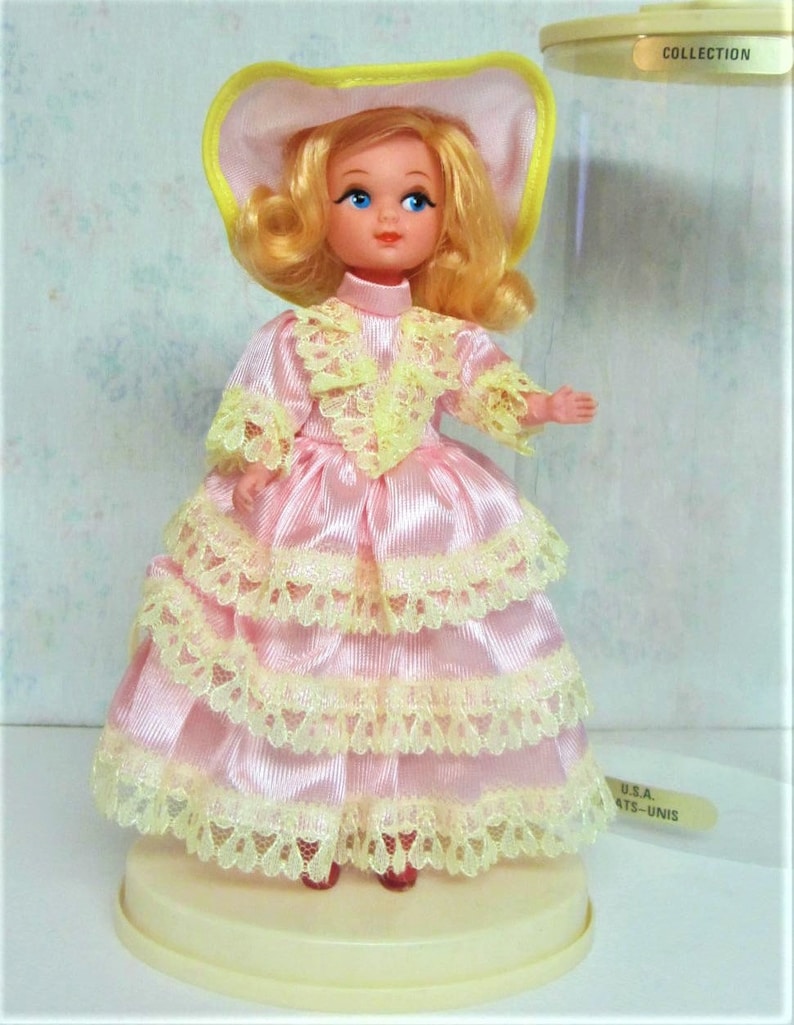 Puede incluir: Una peque&ntilde;a mu&ntilde;eca de porcelana que lleva un vestido rosa con ribetes de encaje amarillo y un sombrero amarillo y blanco. La mu&ntilde;eca est&aacute; de pie sobre una base blanca y se encuentra en una vitrina de pl&aacute;stico transparente. La vitrina tiene una etiqueta que dice "COLLECTION".