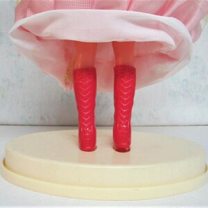 Puede incluir: Una mu&ntilde;eca rosa con botas rojas de pie sobre una base ovalada blanca. La mu&ntilde;eca lleva un vestido rosa con una enagua blanca.