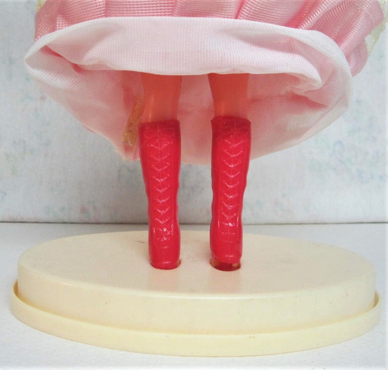 Puede incluir: Una mu&ntilde;eca rosa con botas rojas de pie sobre una base ovalada blanca. La mu&ntilde;eca lleva un vestido rosa con una enagua blanca.
