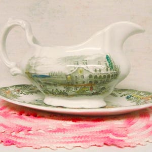 May include: A white ceramic gravy boat with a landscape design, resting on a matching saucer. The boat has a curved handle and spout. The saucer and boat are on a pink and white crocheted doily.