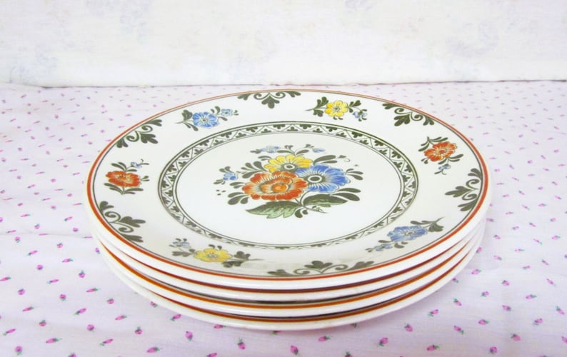 May include: A stack of four white ceramic plates with a floral design in shades of green, yellow, orange, and blue. The plates have a brown rim and a green and white border.