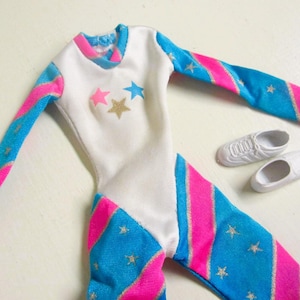 Peut inclure: Une tenue de poupée blanche, bleue et rose avec des étoiles. Le vêtement une pièce a des manches et des jambes rayées bleues et roses, avec des étoiles dorées et roses sur le devant. Comprend une paire de baskets blanches.