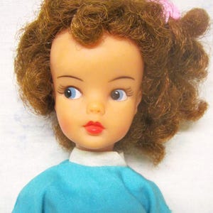 May include: A vintage doll with brown curly hair, blue eyes, and red lips. She is wearing a blue shirt with a white collar.