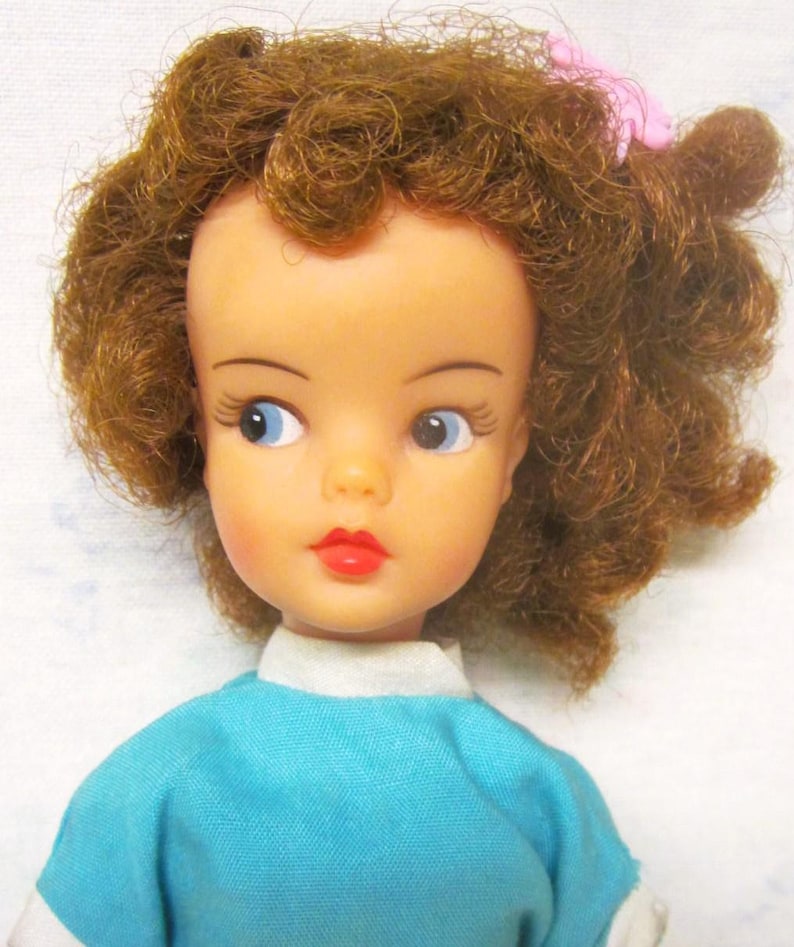 May include: A vintage doll with brown curly hair, blue eyes, and red lips. She is wearing a blue shirt with a white collar.