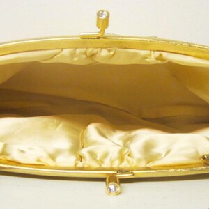 K&ouml;nnte beinhalten: Offene Ansicht einer Vintage-Gold-Clutch. Die Handtasche hat einen goldenen Rahmen und Verschluss mit einem kleinen Strass-Akzent. Der Innenraum ist mit einem hellgelben Satin-Stoff gef&uuml;ttert. Ein goldener Kettenriemen ist sichtbar.