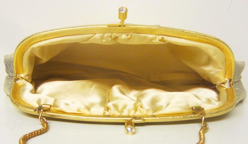 K&ouml;nnte beinhalten: Offene Ansicht einer Vintage-Gold-Clutch. Die Handtasche hat einen goldenen Rahmen und Verschluss mit einem kleinen Strass-Akzent. Der Innenraum ist mit einem hellgelben Satin-Stoff gef&uuml;ttert. Ein goldener Kettenriemen ist sichtbar.