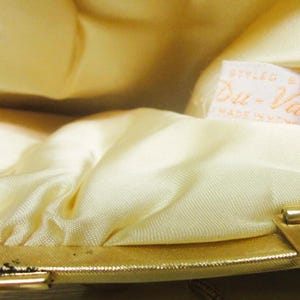 K&ouml;nnte beinhalten: Nahaufnahme des Inneren einer Vintage-Handtasche. Das Futter ist ein weicher, hellgelber Satin, der mit dem goldfarbenen Metallrahmen und -verschluss kontrastiert. Ein Etikett lautet "Styled by Du-Val, Made in Hong Kong."