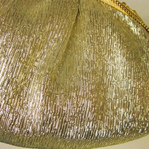 K&ouml;nnte beinhalten: Nahaufnahme eines goldfarbenen, strukturierten Stoffes. Das Material hat ein schimmerndes, metallisches Aussehen mit einem diagonalen Rippenmuster. Eine goldene Kette ist am Rand des Stoffes zu sehen.