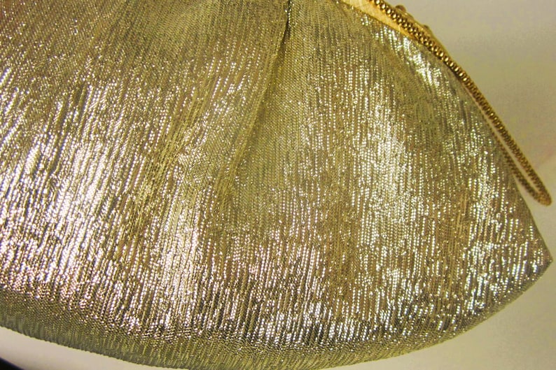 K&ouml;nnte beinhalten: Nahaufnahme eines goldfarbenen, strukturierten Stoffes. Das Material hat ein schimmerndes, metallisches Aussehen mit einem diagonalen Rippenmuster. Eine goldene Kette ist am Rand des Stoffes zu sehen.