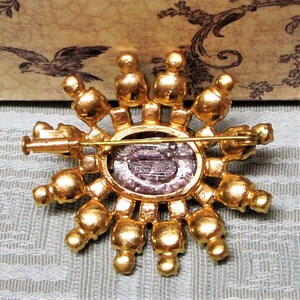 Peut inclure: Une broche dor&eacute;e avec un grand strass ovale violet au centre. La broche est entour&eacute;e de plus petits strass ronds dispos&eacute;s en cercle.