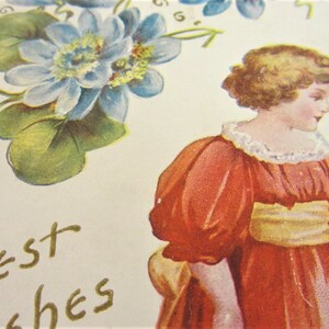 Puede incluir: Una postal vintage con una jovencita con un vestido rojo sosteniendo una peque&ntilde;a caja marr&oacute;n. La ni&ntilde;a tiene el pelo rubio y mira hacia la derecha. La postal tiene un dise&ntilde;o floral con flores azules y hojas verdes. El texto "Mejores deseos" est&aacute; escrito en letras doradas.