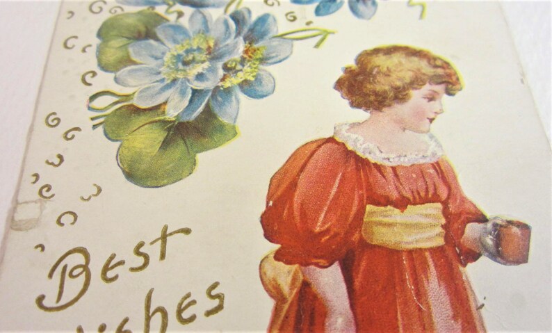 Puede incluir: Una postal vintage con una jovencita con un vestido rojo sosteniendo una peque&ntilde;a caja marr&oacute;n. La ni&ntilde;a tiene el pelo rubio y mira hacia la derecha. La postal tiene un dise&ntilde;o floral con flores azules y hojas verdes. El texto "Mejores deseos" est&aacute; escrito en letras doradas.