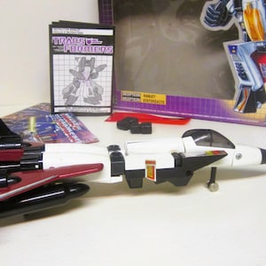 Peut inclure: Un jouet d'avion de chasse blanc et rouge avec des accents noirs et une bande bleue. Le jouet est en mode robot et possède un cockpit. Le jouet fait partie de la ligne de jouets Transformers. La boîte du jouet est visible en arrière-plan.