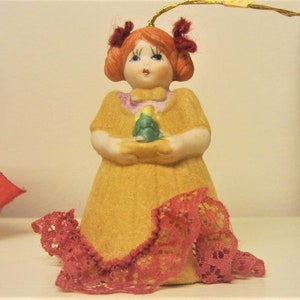 Peut inclure: Une figurine de poupée en porcelaine aux cheveux roux, vêtue d'une robe jaune avec une bordure en dentelle rouge. Elle tient un petit arbre de Noël vert.