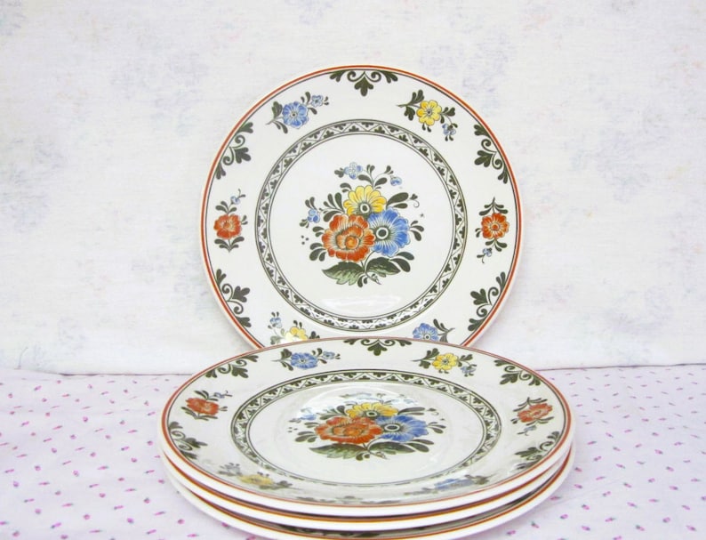 May include: A set of three white ceramic dinner plates with a floral design. The plates feature a green and brown border with a floral pattern in shades of red, yellow, blue, and green.