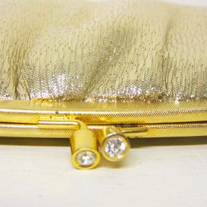 K&ouml;nnte beinhalten: Eine goldene Clutch-Tasche mit einer strukturierten, schimmernden Oberfl&auml;che. Die Tasche hat einen goldenen Rahmen und Verschluss, mit zwei dekorativen, goldfarbenen Verschl&uuml;ssen, die jeweils einen klaren, runden Edelstein aufweisen. Die Tasche ist im Vintage-Stil.