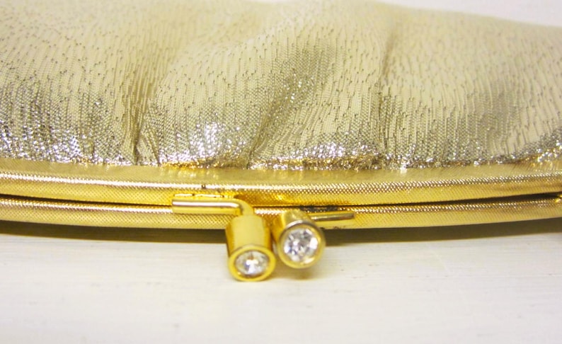 K&ouml;nnte beinhalten: Eine goldene Clutch-Tasche mit einer strukturierten, schimmernden Oberfl&auml;che. Die Tasche hat einen goldenen Rahmen und Verschluss, mit zwei dekorativen, goldfarbenen Verschl&uuml;ssen, die jeweils einen klaren, runden Edelstein aufweisen. Die Tasche ist im Vintage-Stil.