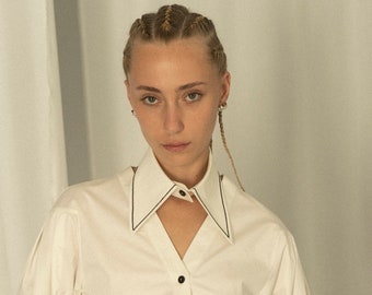 May include: A cream-coloured button-down shirt with a unique collar design. The collar has a black trim and a cutout detail. The shirt has long sleeves and black buttons. The model is wearing black trousers.