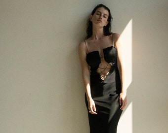 May include: A black, floor-length dress with spaghetti straps and a cut-out design on the bodice. The dress features a fitted silhouette and a subtle sheen. The model is leaning against a white wall, with sunlight casting shadows.