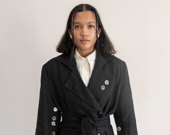 May include: A black linen coat with a wrap belt and large buttons. The coat has a double-breasted design with large pockets and buttons on the sleeves. The model is wearing a white collared shirt underneath.
