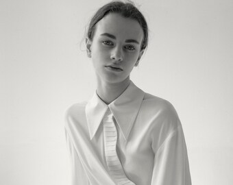 May include: A black and white photograph of a white long-sleeve shirt with a unique pleated design on the front. The shirt is paired with black corduroy pants. The model is looking directly at the camera.