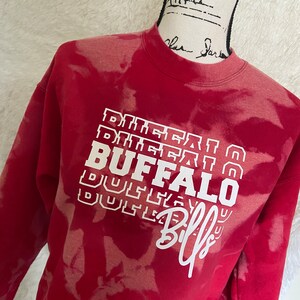 May include: Red sweatshirt with a white bleach tie-dye pattern. The sweatshirt has a white graphic that says "BUFFALO" with the word "Bills" written in cursive underneath.