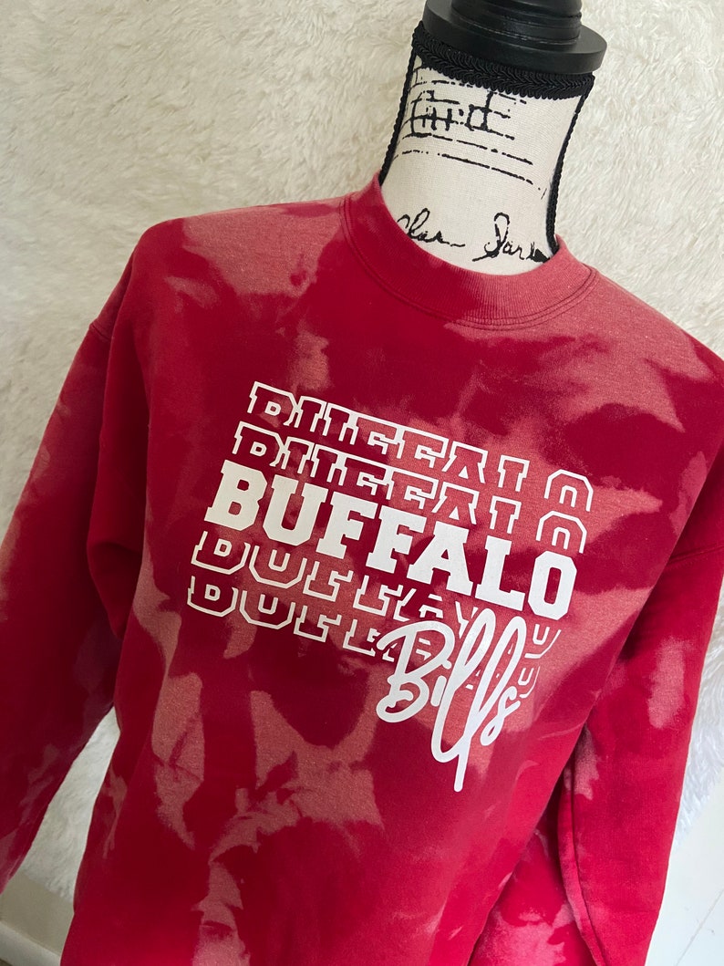 May include: Red sweatshirt with a white bleach tie-dye pattern. The sweatshirt has a white graphic that says "BUFFALO" with the word "Bills" written in cursive underneath.