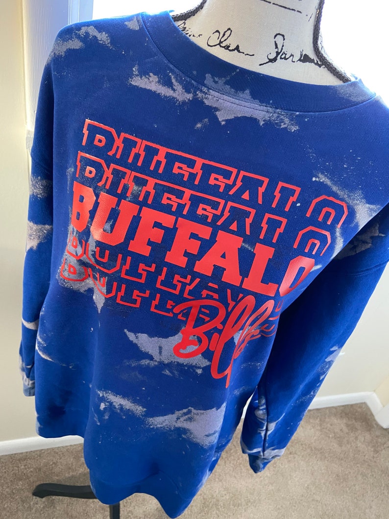 May include: Blue sweatshirt with a white tie-dye pattern and red text that says "Buffalo Bills" in a stacked design.