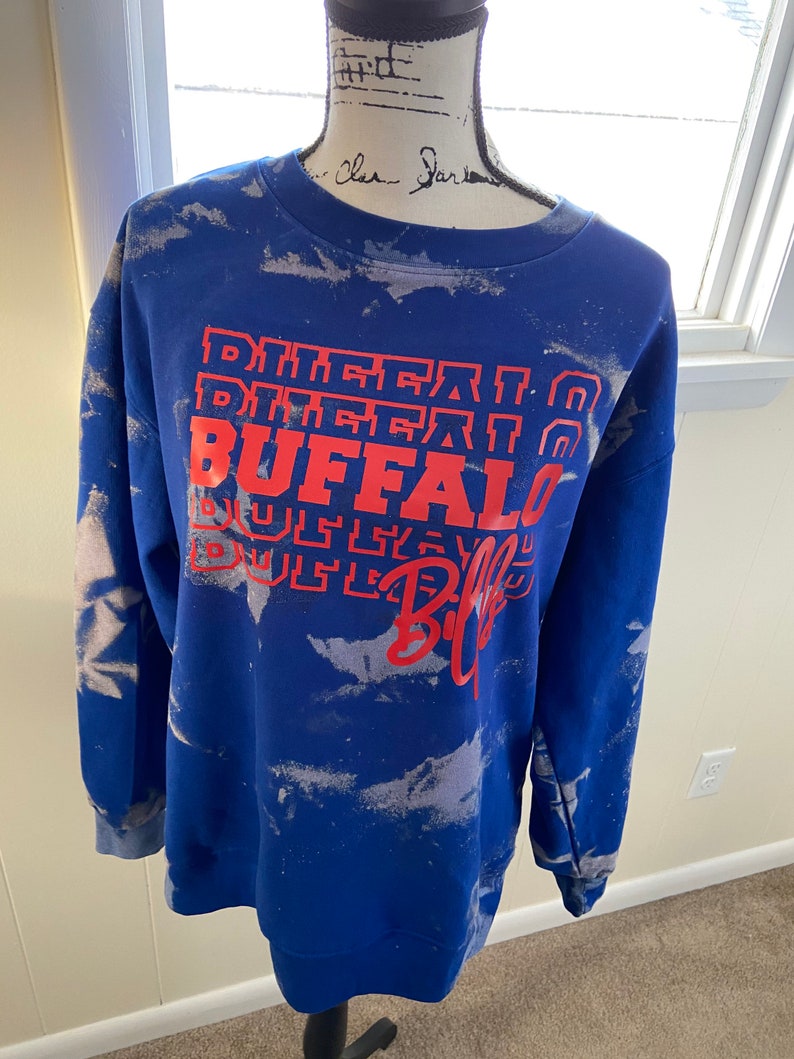 May include: Blue sweatshirt with a white tie-dye pattern and red text that says "Buffalo Bills" in a stacked design.