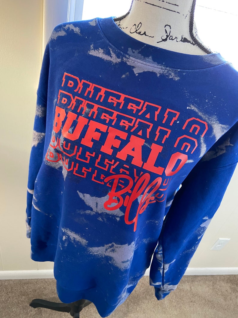 May include: Blue tie-dye sweatshirt with white splatter paint and red text that reads "Buffalo Bills" in a layered design.