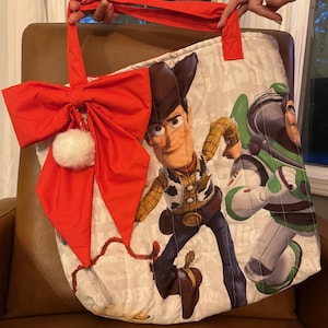 Pode incluir: Uma sacola tote acolchoada com os personagens Woody e Buzz Lightyear de Toy Story. A sacola tem um la&ccedil;o vermelho com um pompom branco e al&ccedil;as vermelhas. A sacola &eacute; segurada por uma pessoa.