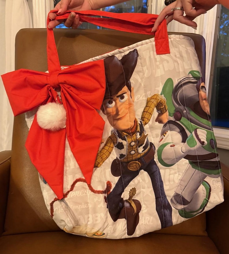 Pode incluir: Uma sacola tote acolchoada com os personagens Woody e Buzz Lightyear de Toy Story. A sacola tem um la&ccedil;o vermelho com um pompom branco e al&ccedil;as vermelhas. A sacola &eacute; segurada por uma pessoa.