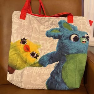 Pode incluir: Uma sacola tote acolchoada com personagens de desenhos animados. A sacola tem al&ccedil;as vermelhas e um fundo bege claro com texto impresso. A frente exibe um pintinho amarelo e um personagem de coelho azul. A sacola &eacute; projetada com um padr&atilde;o acolchoado.