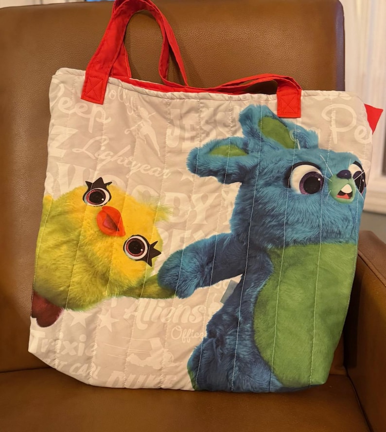 Pode incluir: Uma sacola tote acolchoada com personagens de desenhos animados. A sacola tem al&ccedil;as vermelhas e um fundo bege claro com texto impresso. A frente exibe um pintinho amarelo e um personagem de coelho azul. A sacola &eacute; projetada com um padr&atilde;o acolchoado.
