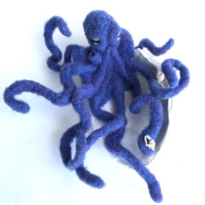 May include: A handcrafted, blue felt octopus with curled tentacles. The octopus is positioned on a silver-colored, shell-shaped base. The felt has a textured appearance, and the octopus has small, light-colored eyes.