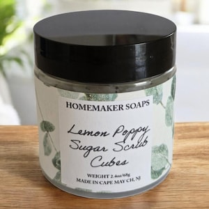 May include: A clear glass jar with a black lid, labeled "HOMEMAKER SOAPS Lemon Poppy Sugar Scrub Cubes." The label features a botanical design. The jar contains a green scrub and weighs 2.4 oz/68g.