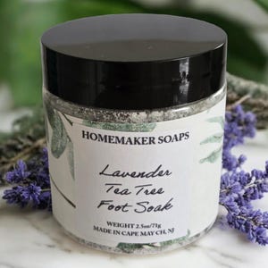 May include: A glass jar with a black lid labeled "HOMEMAKER SOAPS Lavender Tea Tree Foot Soak." The jar contains a foot soak product, with a weight of 2.5 oz (71g). The jar is surrounded by purple lavender flowers.