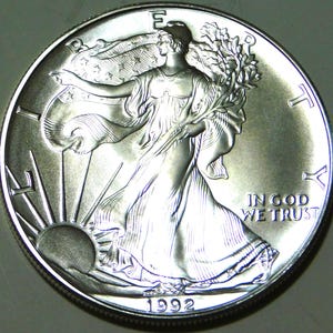 May include: A silver coin featuring Lady Liberty walking towards the sun with an American flag draped over her shoulders. The coin has the inscription "IN GOD WE TRUST" and the year "1992".