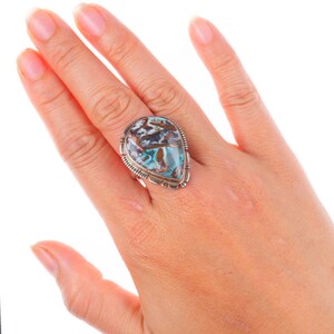 May include: A silver ring featuring a large, teardrop-shaped stone with a unique pattern of blue, brown, and white hues. The ring has a detailed silver setting with a textured border, showcasing a Southwestern style.