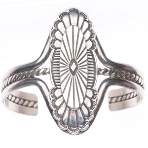 May include: A silver cuff bracelet featuring a large, elongated oval centerpiece with radiating sunburst lines and a central diamond-shaped cutout. The band is constructed from two smooth silver wires flanking a central twisted rope wire.